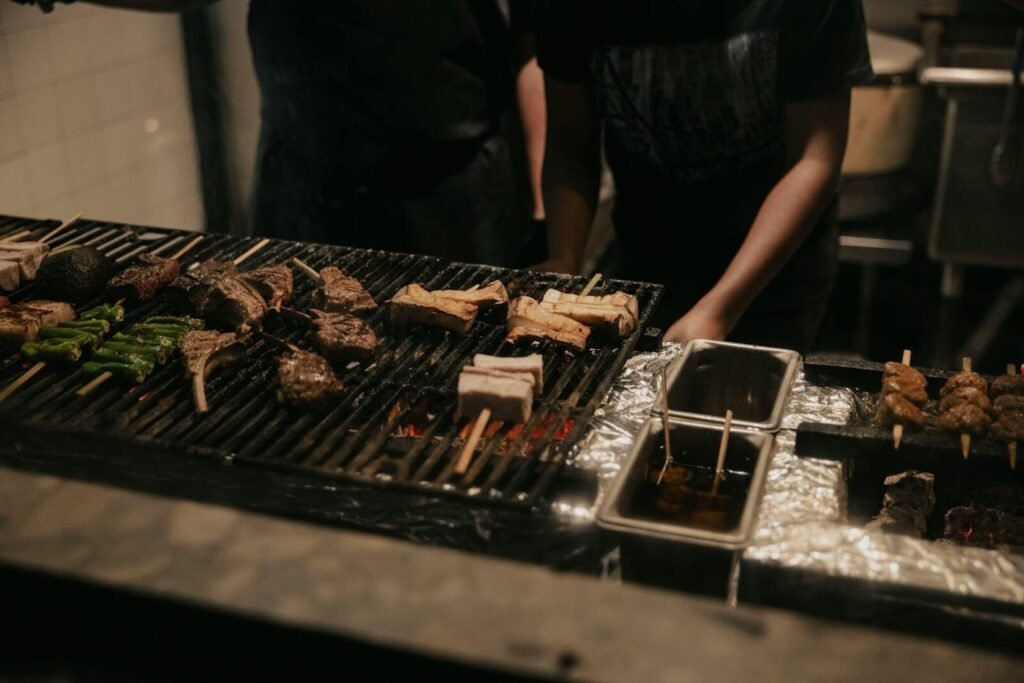 Close-up of skewers being grilled over open flame by people, featuring various meats and vegetables.